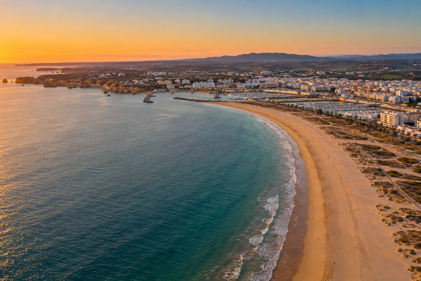 Aerial view of a long sandy beach curving toward a town and a harbor jetty, with the sun low over the sea (illustration)